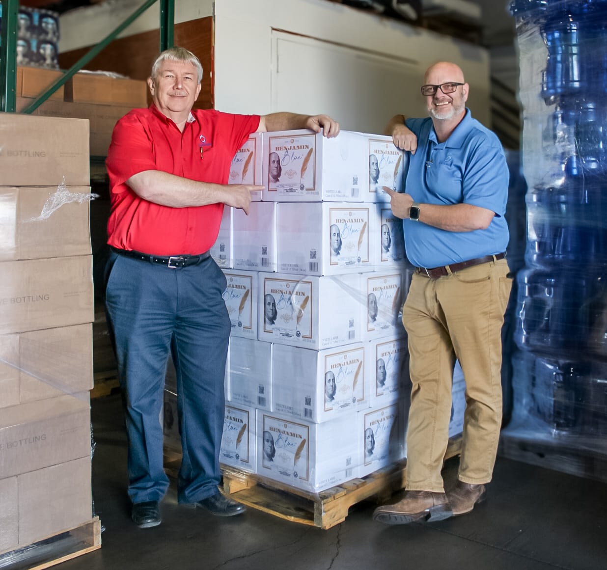 Two workers for Texas Water House, one in a blue shirt and one in a red shirt, standing by a pallet of their water products.