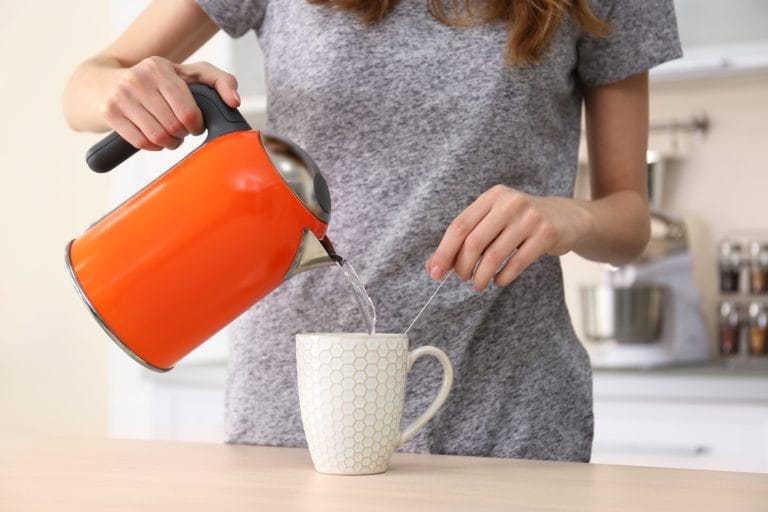 A woman in a gray shirt pouring hot water into a tea cup from an orange kettle.